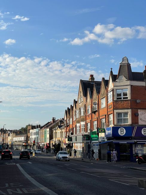 A street scene showing a row of multi-storey residential buildings with brick and stucco facades, pitched roofs, and large windows, situated along a busy urban pavement under a blue sky with scattered